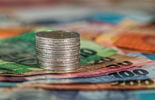 Stos srebrnych monet na tle rozmazanych kolorowych banknotów różnych walut.