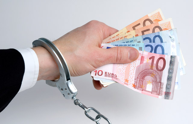 Ręka w kajdankach trzymająca banknoty euro różnych nominałów.